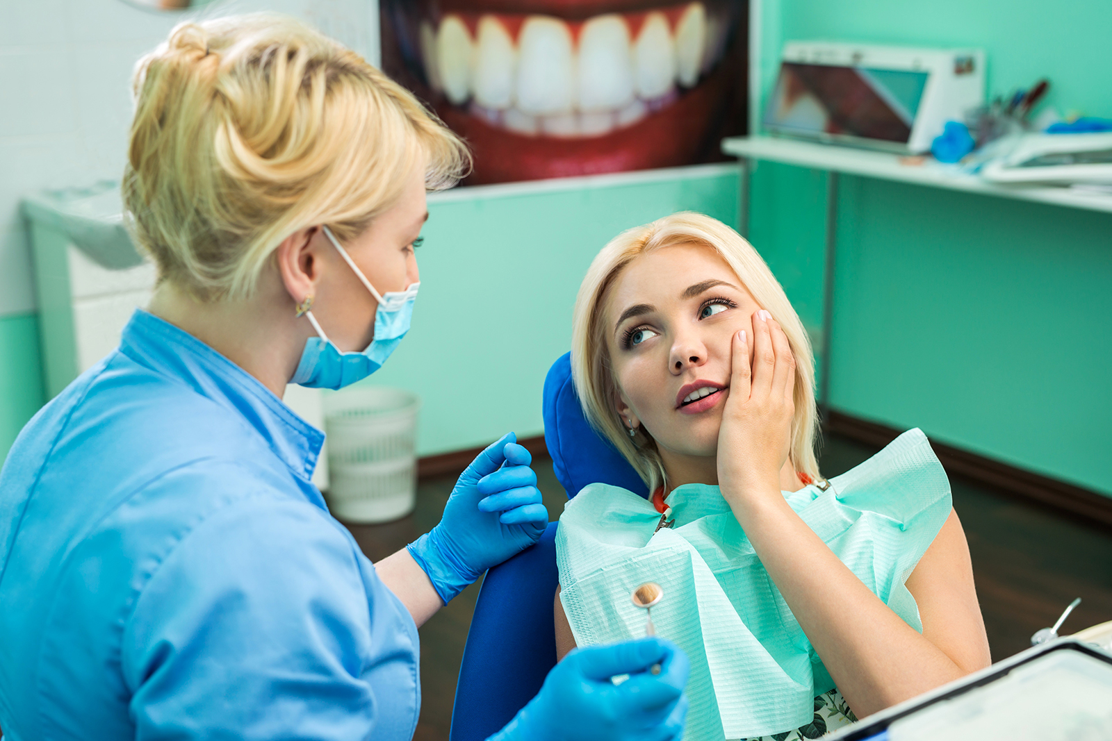 The image depicts a dental office setting where a female dentist is examining a woman s teeth while she sits in the chair, both wearing masks.