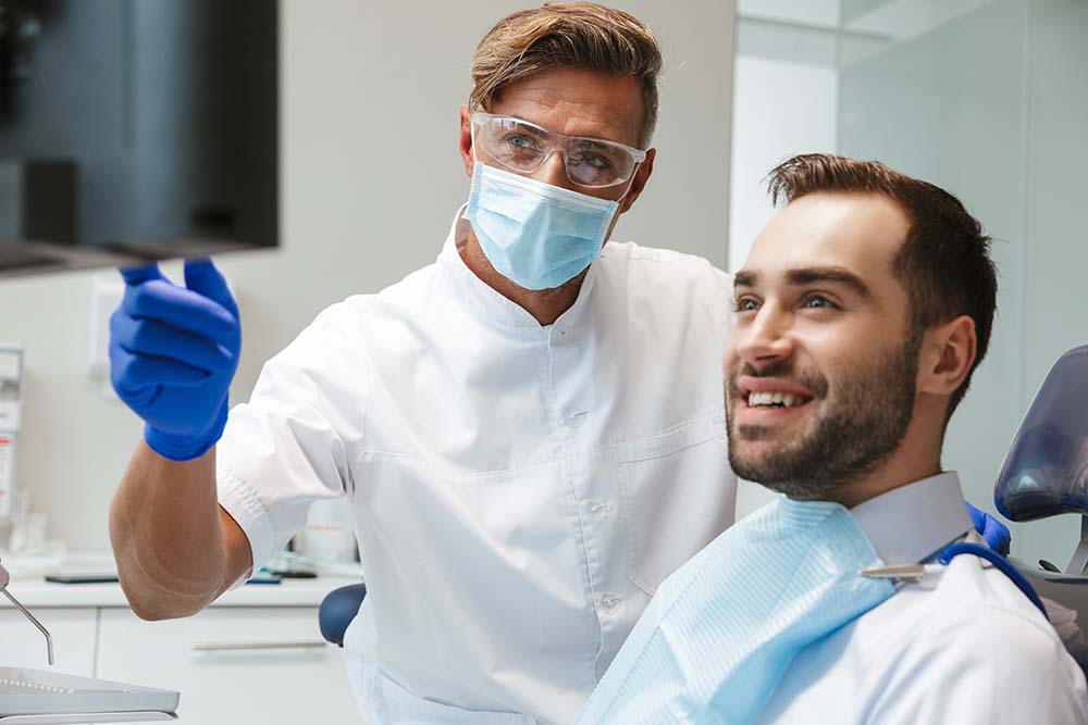 A dental professional in protective gear pointing at a man s mouth with a mirror while he is seated in a dentist s chair, both smiling.