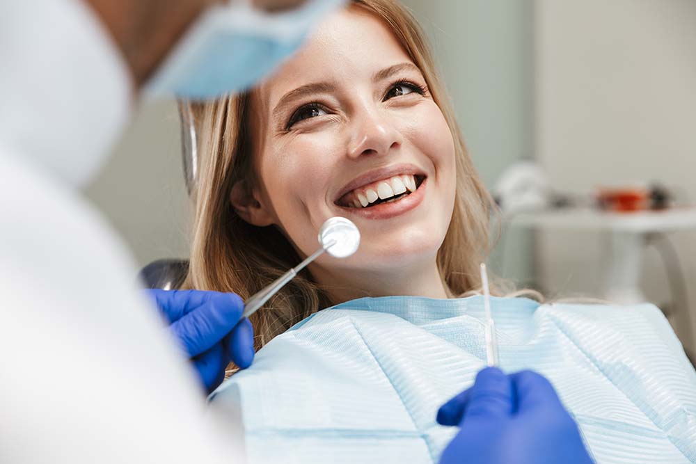 A woman smiling while sitting in a dental chair with a dentist performing a procedure behind her.
