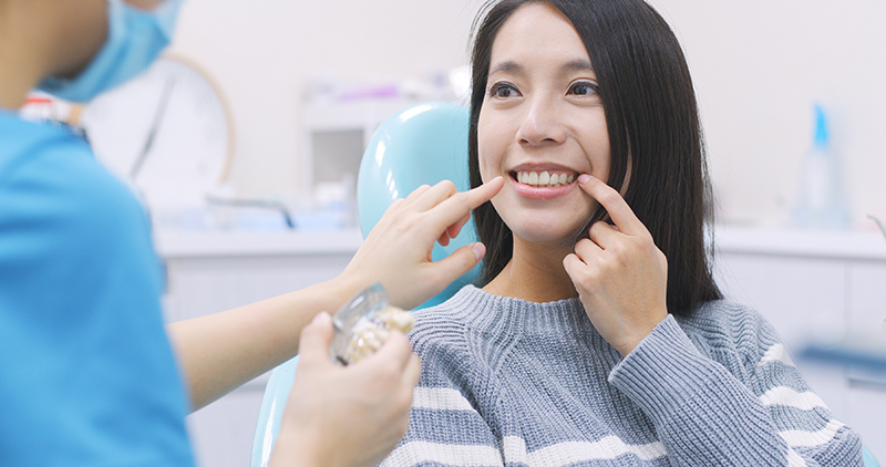A woman sitting in a dental chair with a dental hygienist attending to her, smiling at the camera while receiving dental care.