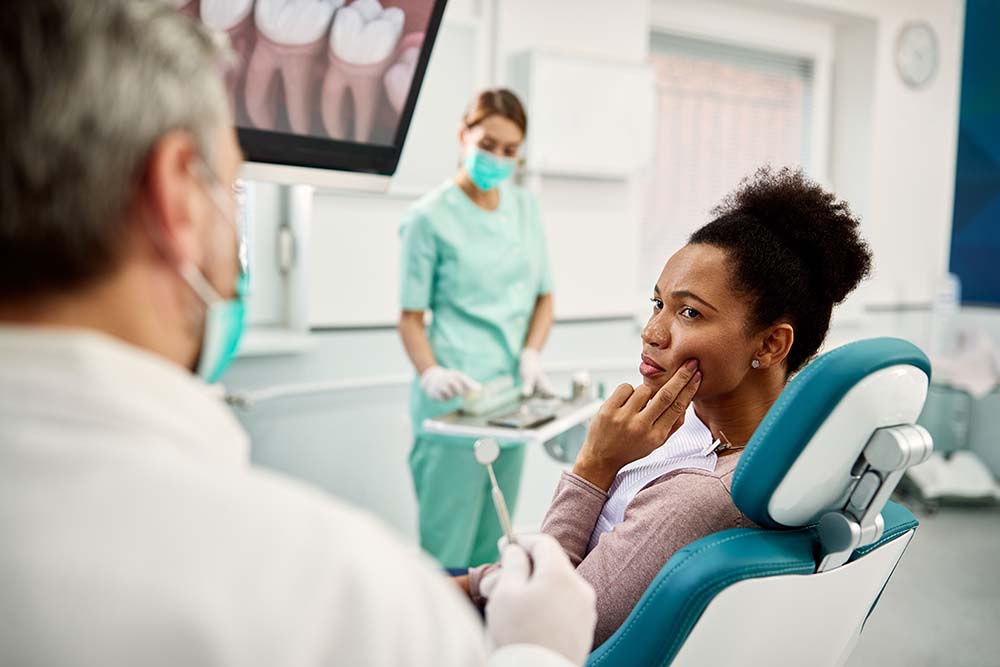 A woman sitting in a dental chair with her hand on her face, receiving dental care from a dentist assisted by another person wearing protective eyewear.